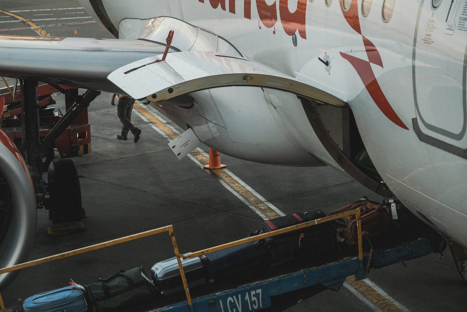 A close-up shot of the side of an airplane, with luggage being loaded onto it from a conveyor belt. The airplane has a white body with red and white markings, and the conveyor belt is blue and yellow. The ground is dark gray.