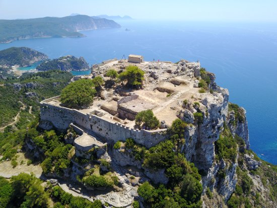  An aerial view of Angelokastro, a Byzantine fortress perched on a rocky cliff overlooking the Ionian Sea in Corfu, Greece. The fortress walls and ruins are visible on the summit, surrounded by lush green vegetation. The deep blue sea stretches out to the horizon, with distant islands visible.