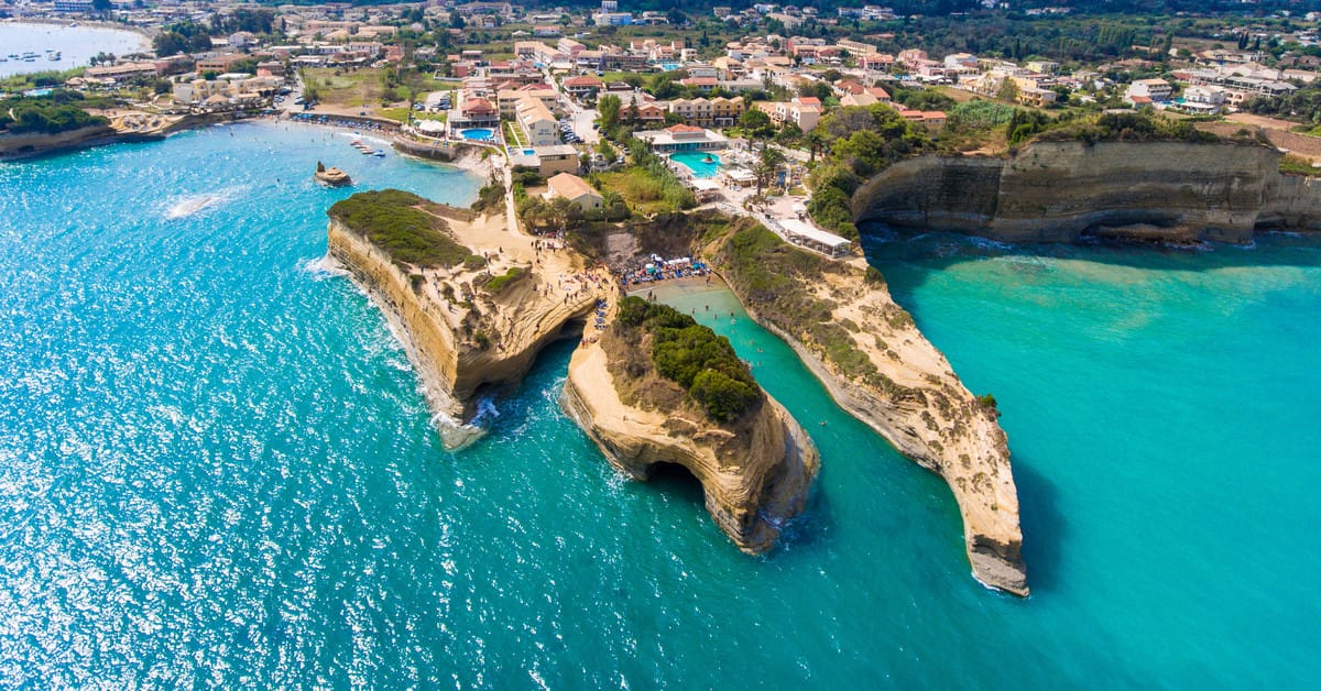 An aerial view of Canal d'Amour in Sidari, Corfu, Greece, showcasing unique sandstone cliffs extending into the turquoise sea. The cliffs create small coves and inlets, with people visible on the beach. A town with buildings and a swimming pool is visible in the background. The water is a vibrant turquoise, and the sky is clear.