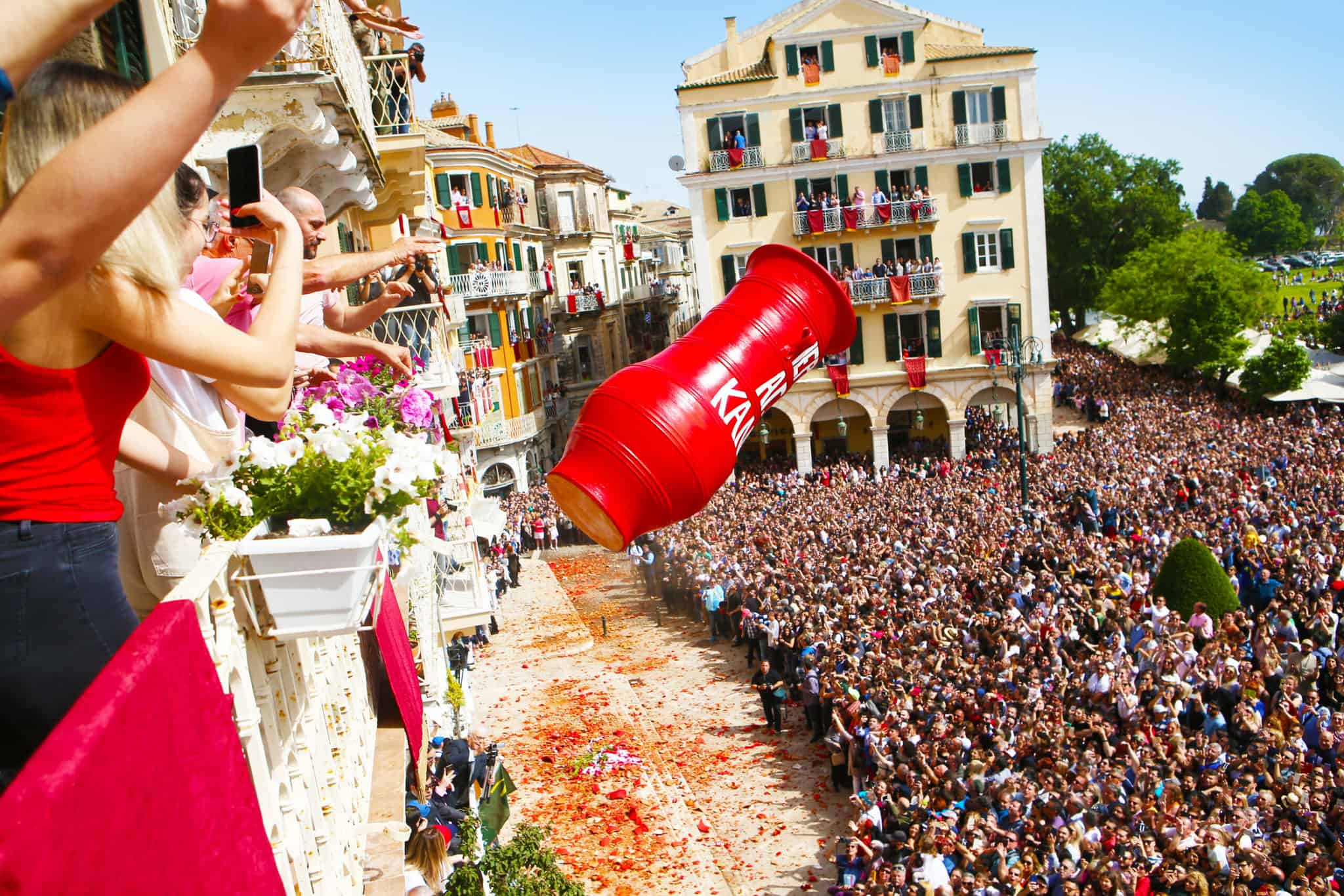 A large red clay pot, known as a "botis," is being thrown from a balcony into a crowd of people gathered on a street in Corfu, Greece, during the Easter Botides custom. People on the balcony are taking photos and videos, while the crowd below eagerly anticipates the smashing of the pot. The street is scattered with red flower petals and broken pieces of clay. The buildings surrounding the street have balconies filled with spectators.