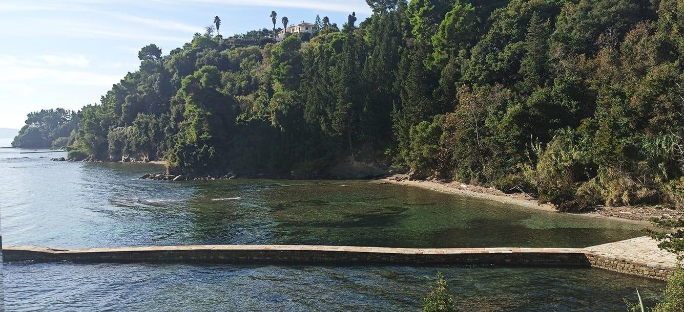 A scenic coastal view in Corfu, Greece, featuring a stone pier extending into calm, clear water. Lush green trees cover a hillside that slopes down to the shoreline. A building is visible on the hilltop in the distance. The sky is partly cloudy. The image captures the natural beauty of the Corfu coastline.