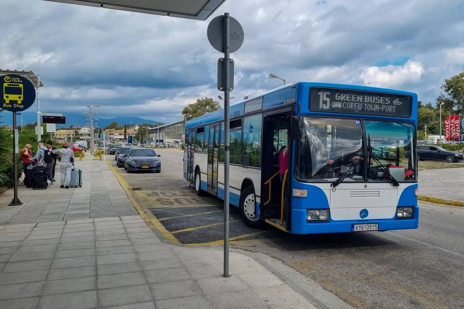 A blue and white Mercedes-Benz O 405 N city bus, labeled '15 GREEN BUSES CORFU TOWN - PORT', is parked at a bus stop in Corfu, Greece. Passengers with luggage are boarding. A 'Bus Stop' sign is visible on the left, and a street sign with a pole is in the center of the image. The license plate of the bus reads 'KYK 3915'. The background shows a cloudy sky, a street with parked cars, and trees. The image captures the scene in Peristeri, Decentralized Administration of Attica, Greece.