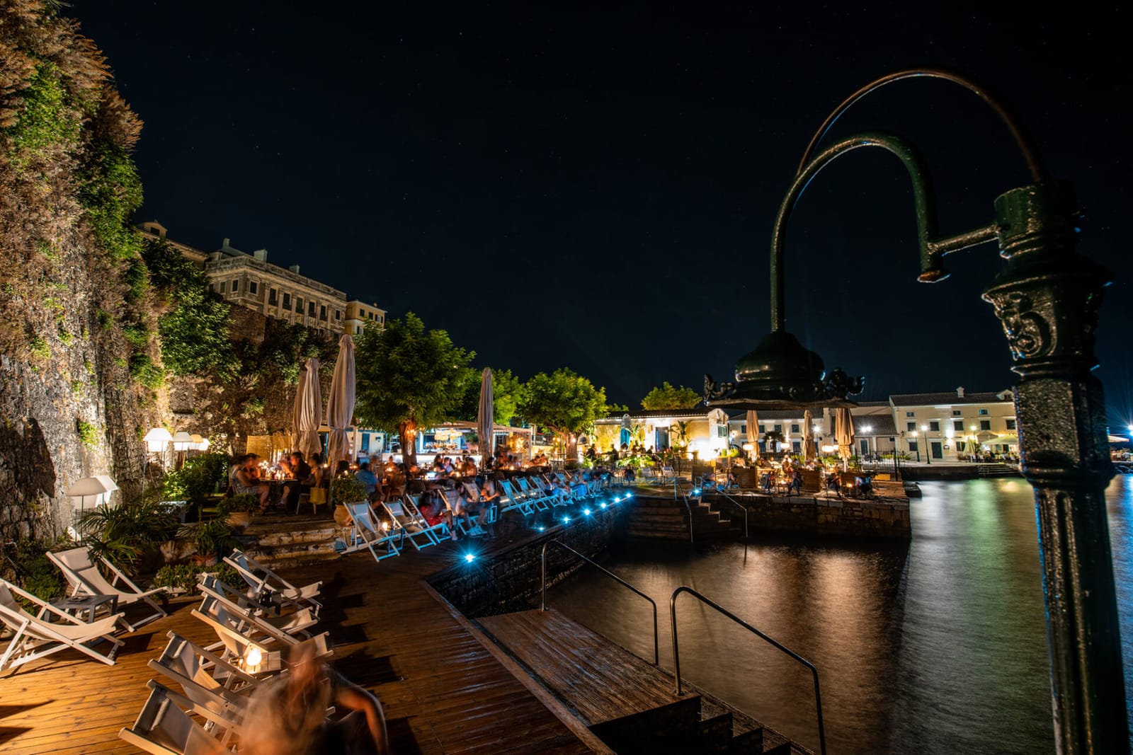 A nighttime scene of a waterfront dining area in Corfu, Greece. The area is illuminated with warm lights, showing tables and chairs set for dining along a wooden boardwalk next to the water. A decorative lamppost is prominent in the foreground. Lounge chairs are neatly arranged along the boardwalk. The water is dark and reflective, and a building is visible on the cliffside in the background. The sky is dark with a few stars visible. The image captures a serene and inviting atmosphere of a night out by the water.