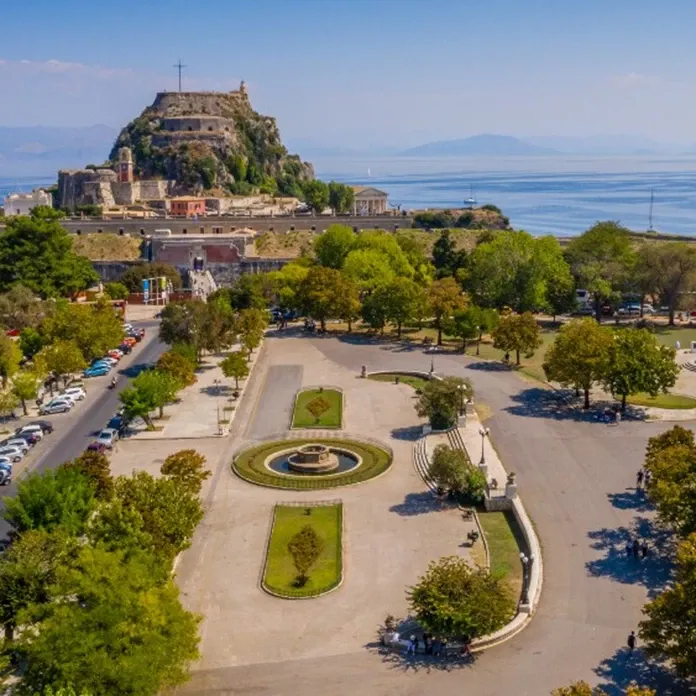 An aerial view of the Spianada Square in Corfu, Greece, with the Old Fortress visible in the background on a rocky hill overlooking the sea. The square features a central circular fountain, paved walkways, and green lawns with trees. The sea is visible in the distance, and cars are parked along one side of the square.