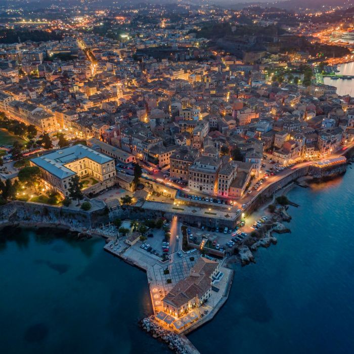 An aerial view of Corfu Town at night, showcasing the city lights illuminating the buildings and streets. The town is nestled along the coastline, with the dark blue sea surrounding it. A prominent building with a courtyard is visible on the left, and a pier extends into the water. The lights create a warm and inviting atmosphere, highlighting the urban landscape. The sky is dark, suggesting twilight or night.
