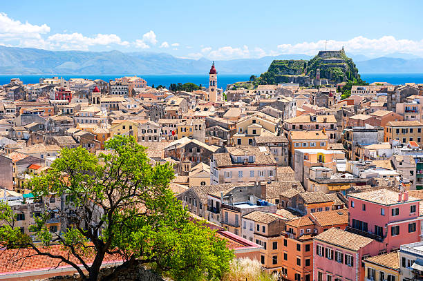 A scenic view of Corfu Town, Greece, showing the densely packed buildings with terracotta roofs stretching towards the coastline. The Old Fortress is visible on a hilltop in the background. A large green tree is in the foreground, framing the cityscape. The blue sea and distant mountains are visible in the background under a bright blue sky with white clouds. The image captures the charm and historical architecture of Corfu.