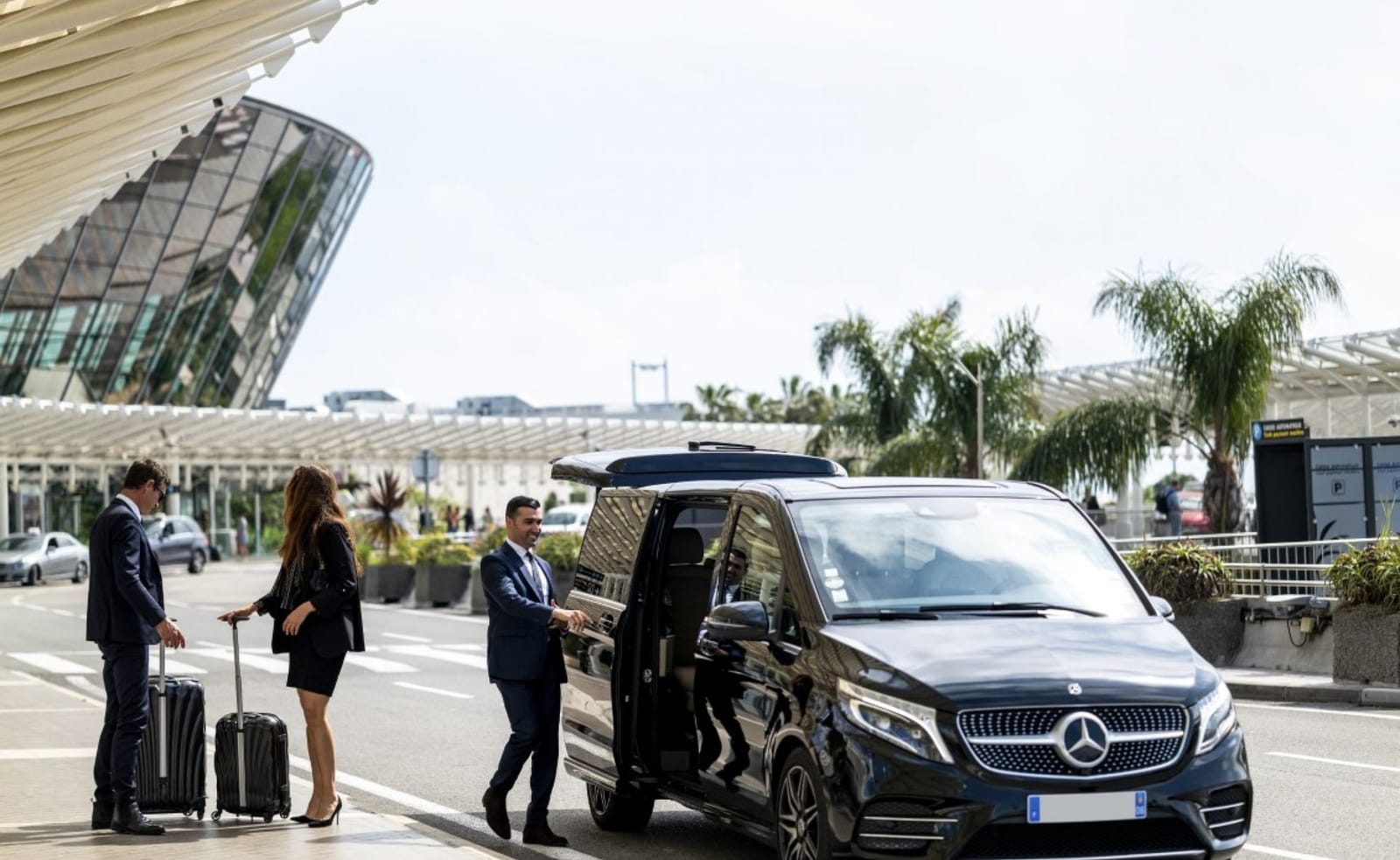A black Mercedes-Benz V-Class minivan is parked at an airport, with three people standing beside it. Two are dressed in suits, one male and one female, each with a rolling suitcase. The third person, a man in a suit, is holding the sliding door open. The airport terminal, featuring a modern glass facade, is visible in the background along with palm trees and other greenery. The scene suggests a luxury airport transfer service.