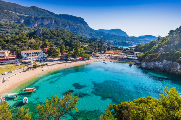 A scenic view of Paleokastritsa Bay in Corfu, Greece, showcasing a turquoise cove surrounded by lush green hills and a sandy beach. Several boats are moored in the clear water, and buildings are visible along the shoreline. The sky is blue and clear.