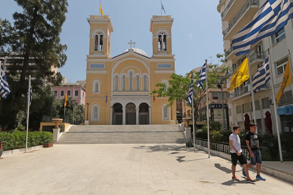 Saint Spyridon Church in Piraeus, Greece, with a yellow facade, twin bell towers, and a wide staircase leading to the entrance. Greek flags are displayed along the street, and two young men are walking in the foreground.