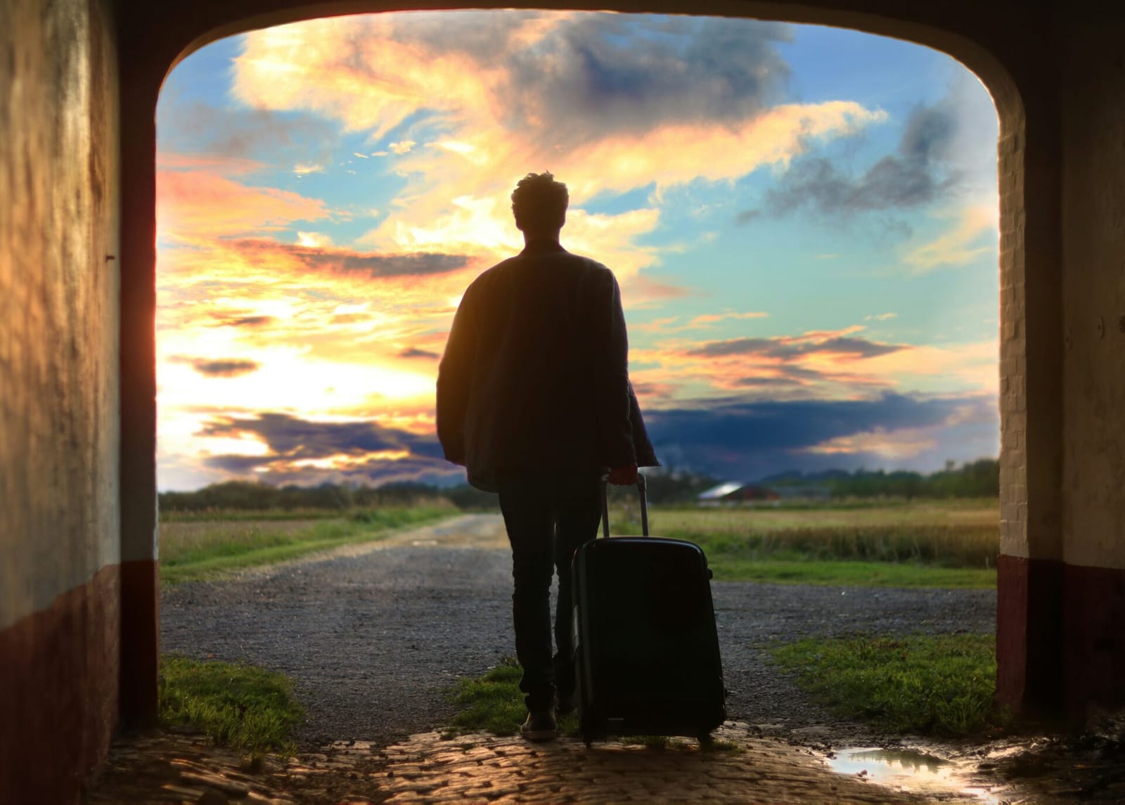 A silhouetted figure of a person standing in an archway with a suitcase, facing a sunset sky and a path leading into a field.