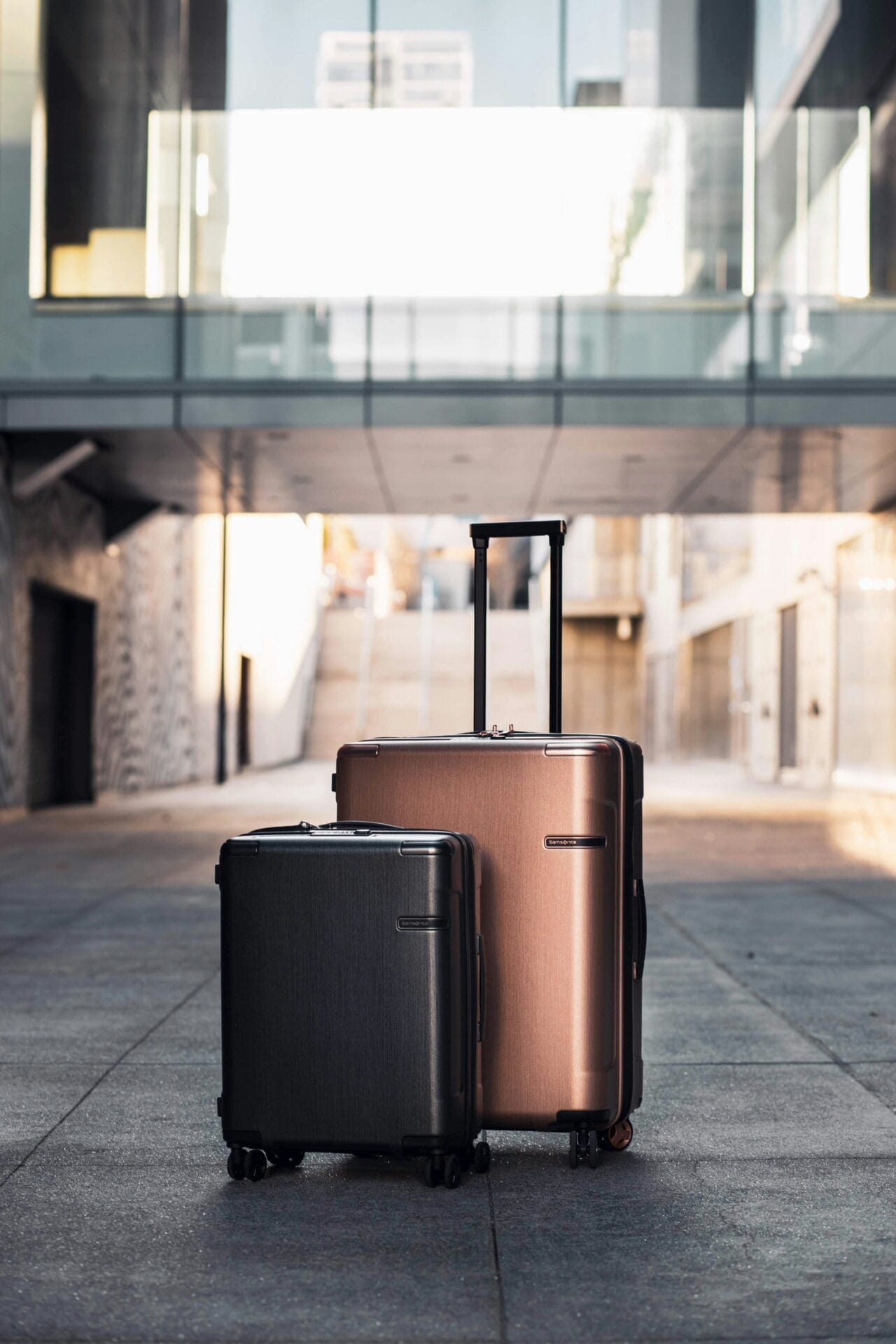 Two suitcases, one dark gray and one bronze, stand side-by-side in an urban setting with modern architecture in the background.